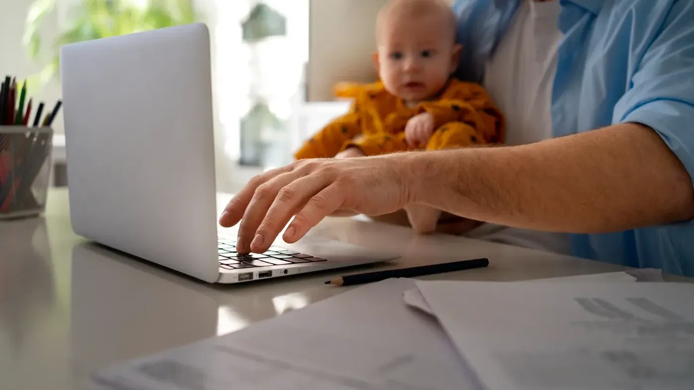 Close-up of a father working on a laptop at a home desk while holding a baby in an orange outfit, representing the ease of insurance online enabling work-life balance and home-based business.