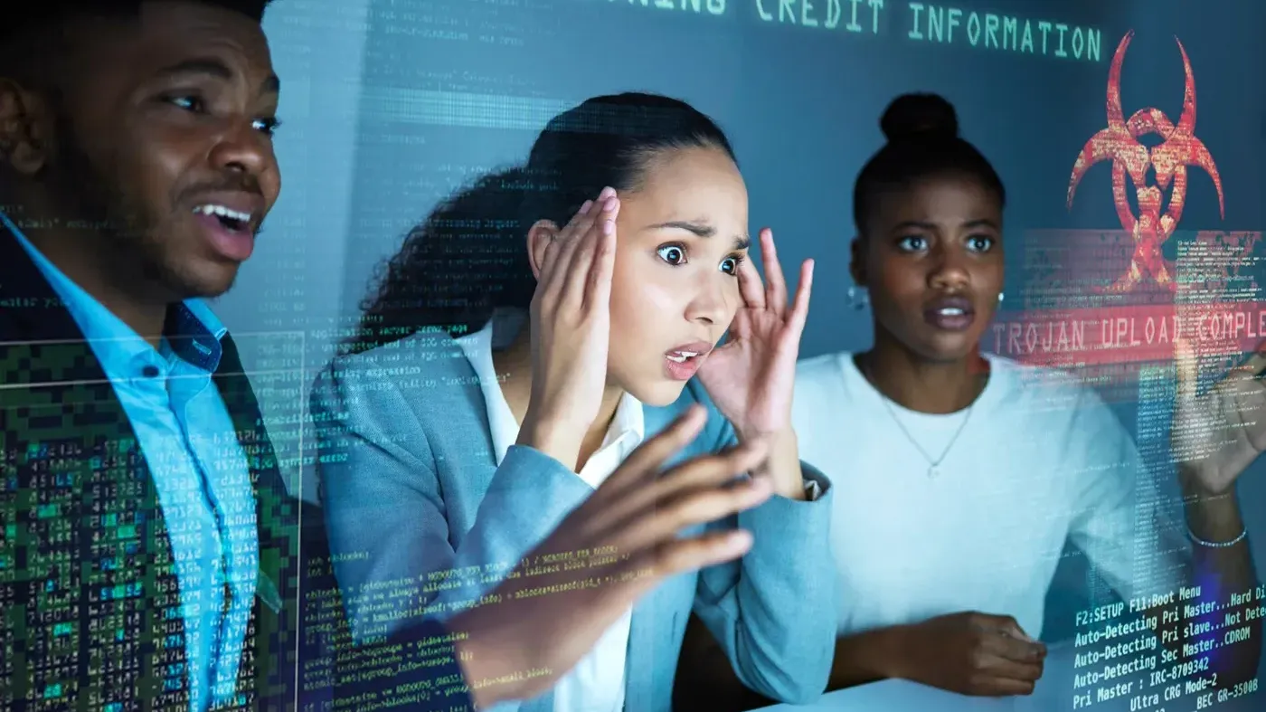 Three diverse business professionals with worried and shocked expressions looking at a screen displaying a red Trojan Upload Complete warning and Cloning Credit Information text during a simulated cyberattack.