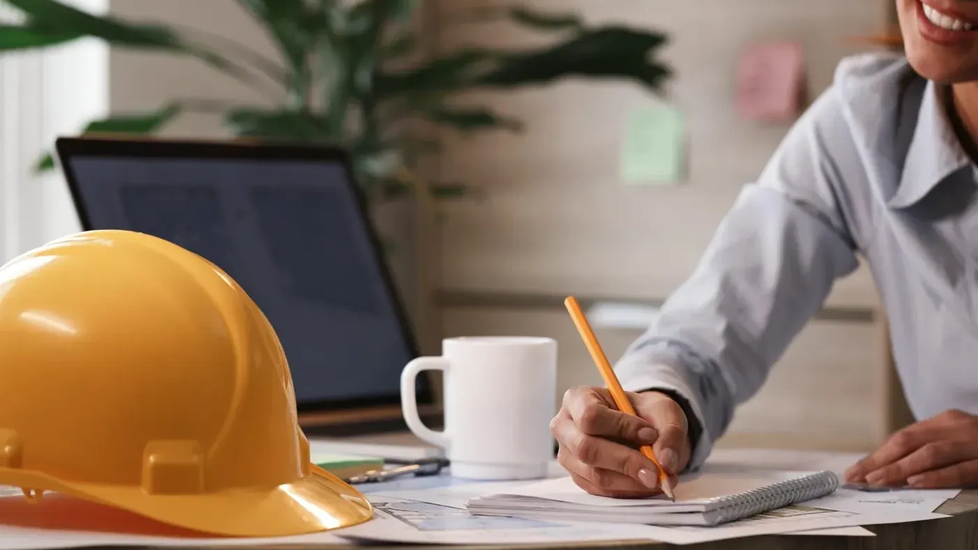 Architect insurance liability professional represented by a smiling female architect writing notes in a notebook at a desk next to a yellow safety helmet and building plans.